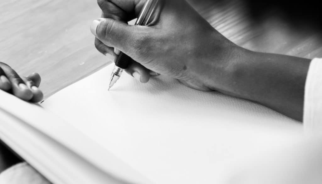 A black and white close-up shot of a person's hand writing in a notebook with a pen.