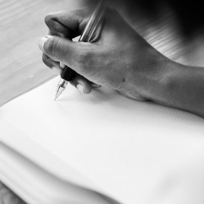 A black and white close-up shot of a person's hand writing in a notebook with a pen.