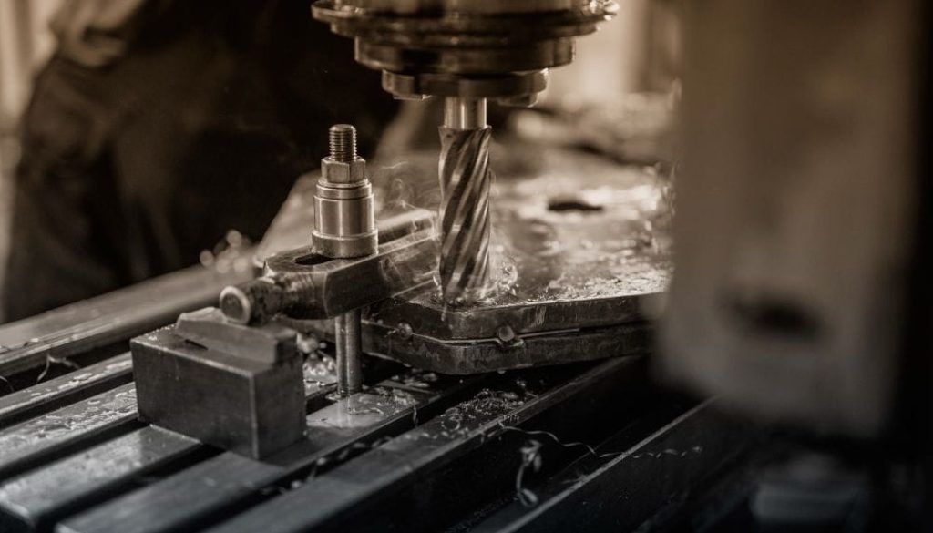 A close-up, high-angle, black-and-white photo of a drill press in a workshop. A spiral drill bit, in focus, cuts into a metal block, creating sparks and metal shavings. The drill head and other machinery are visible in the background.