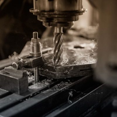 A close-up, high-angle, black-and-white photo of a drill press in a workshop. A spiral drill bit, in focus, cuts into a metal block, creating sparks and metal shavings. The drill head and other machinery are visible in the background.