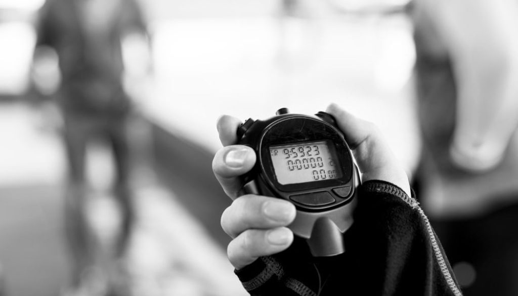 Black and white close-up of a hand holding a digital stopwatch, with the time reading 0:59.23. The background is blurred, showing a figure in motion.