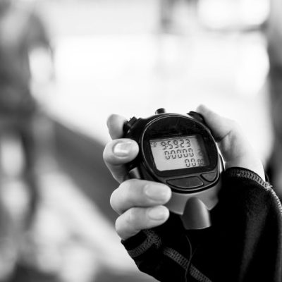 Black and white close-up of a hand holding a digital stopwatch, with the time reading 0:59.23. The background is blurred, showing a figure in motion.