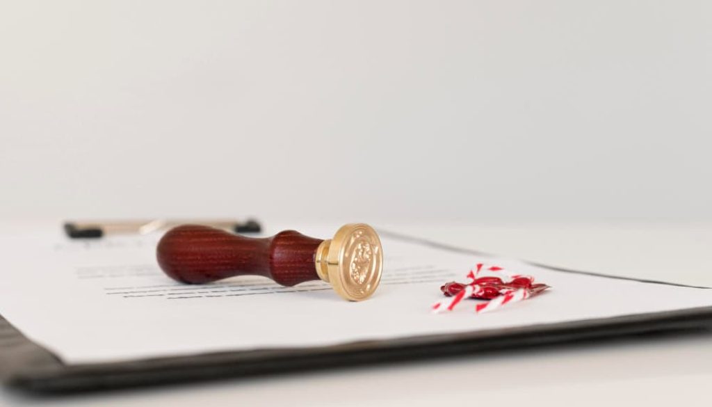 A wooden wax seal stamp and a red wax seal with ribbons rest on a document clipped to a black board.