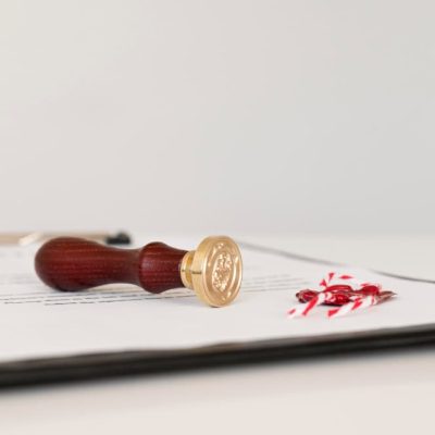 A wooden wax seal stamp and a red wax seal with ribbons rest on a document clipped to a black board.