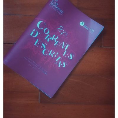 An overhead shot shows a purple program for the 27th edition of "Correntes d’Escritas". The booklet sits on a dark, polished wooden floor with visible planks. The text on the cover indicates the event took place from February 21 to 28, 2026, in Póvoa de Varzim. The title "Correntes d’Escritas" appears in large, stylized teal and white letters. A quote by Álvaro Laborinho Lúcio at the bottom mentions "A Casa".