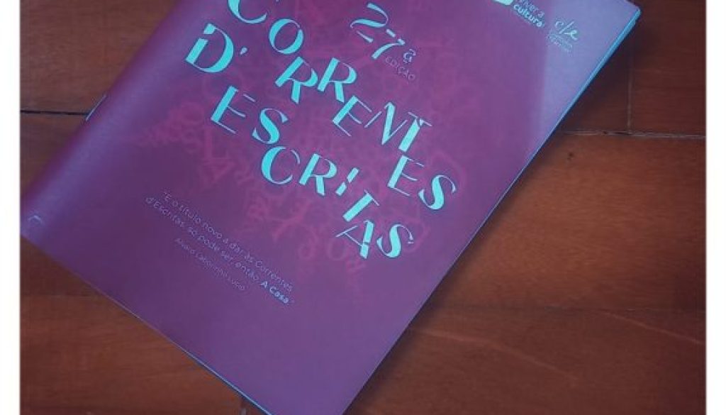 A high-angle, vertical shot shows the official program for the 27th edition of "Correntes d’Escritas" resting diagonally on polished wooden floor. The program's cover is a deep magenta or plum colour, featuring the event title in stylized, light-blue serif lettering. At the top left, the text "2026 21-28 FEVEREIRO PÓVOA DE VARZIM" is printed in white.