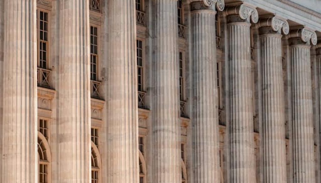 Tall, neoclassical stone columns dominate this side-angle view of a grand building. These fluted pillars feature scroll-like volutes at the top, a hallmark of the Ionic order. The warm, sandy-colored stone glows under direct sunlight, casting deep shadows into the vertical grooves of each column. Behind the rhythmic row of pillars, the building facade reveals multiple levels of tall, narrow windows. Small, decorative metal railings line the window ledges, adding a delicate texture against the massive stone structure. The perspective creates a sense of depth, with the columns receding into the distance and emphasizing the scale of the architecture.