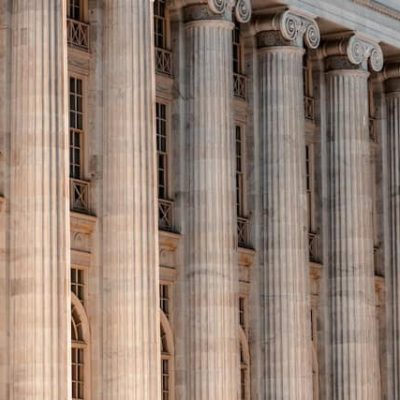 Tall, neoclassical stone columns dominate this side-angle view of a grand building. These fluted pillars feature scroll-like volutes at the top, a hallmark of the Ionic order. The warm, sandy-colored stone glows under direct sunlight, casting deep shadows into the vertical grooves of each column. Behind the rhythmic row of pillars, the building facade reveals multiple levels of tall, narrow windows. Small, decorative metal railings line the window ledges, adding a delicate texture against the massive stone structure. The perspective creates a sense of depth, with the columns receding into the distance and emphasizing the scale of the architecture.